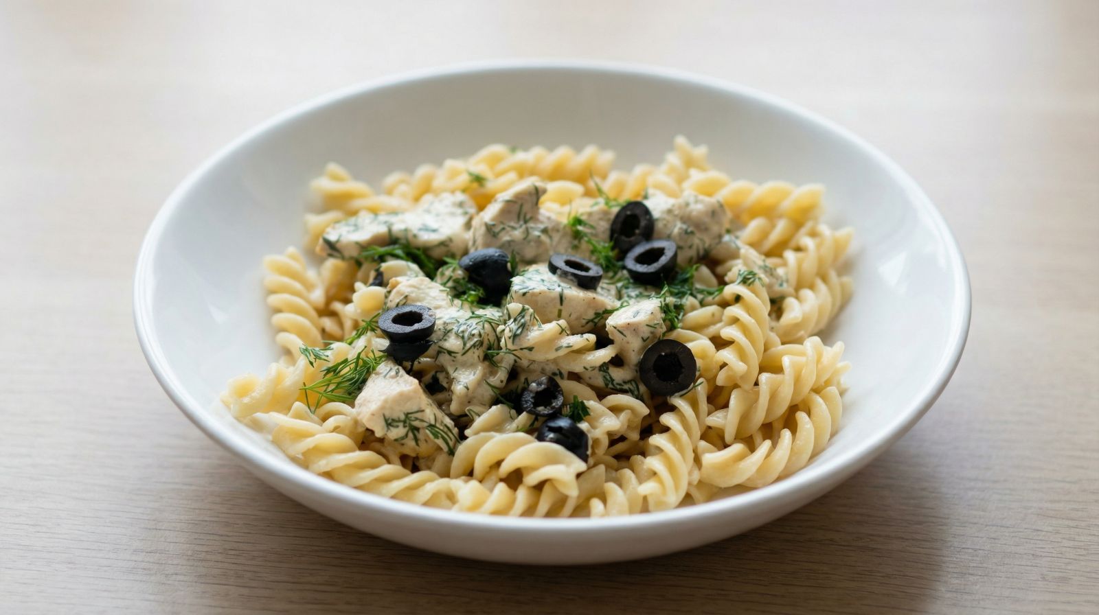 Makaron z kurczakiem w śmietankowym sosie podany na białym talerzu, udekorowany świeżym koperkiem i plasterkami oliwek.