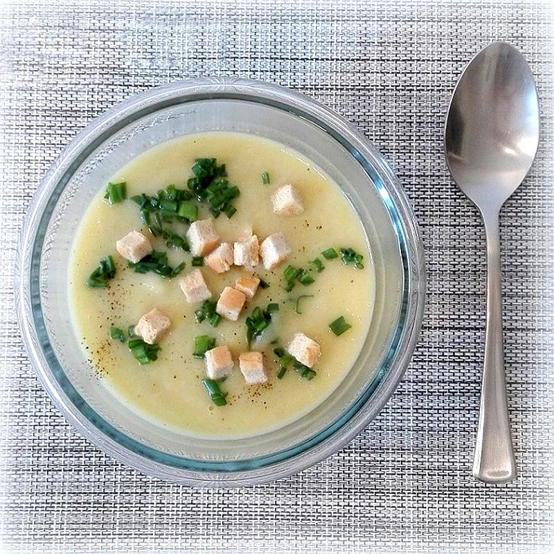 Porowo-ziemniaczana zupa krem w szklanej misce, udekorowana grzankami i posiekanym szczypiorkiem, obok miski leży łyżka.
