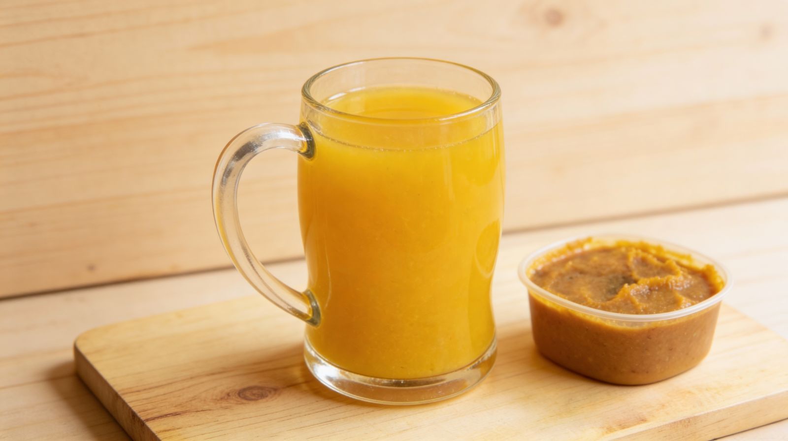 Domowa pasta warzywna w plastikowym pojemniku obok szklanki z rozrobioną bulionetką warzywną — naturalna baza smakowa do zup i sosów.