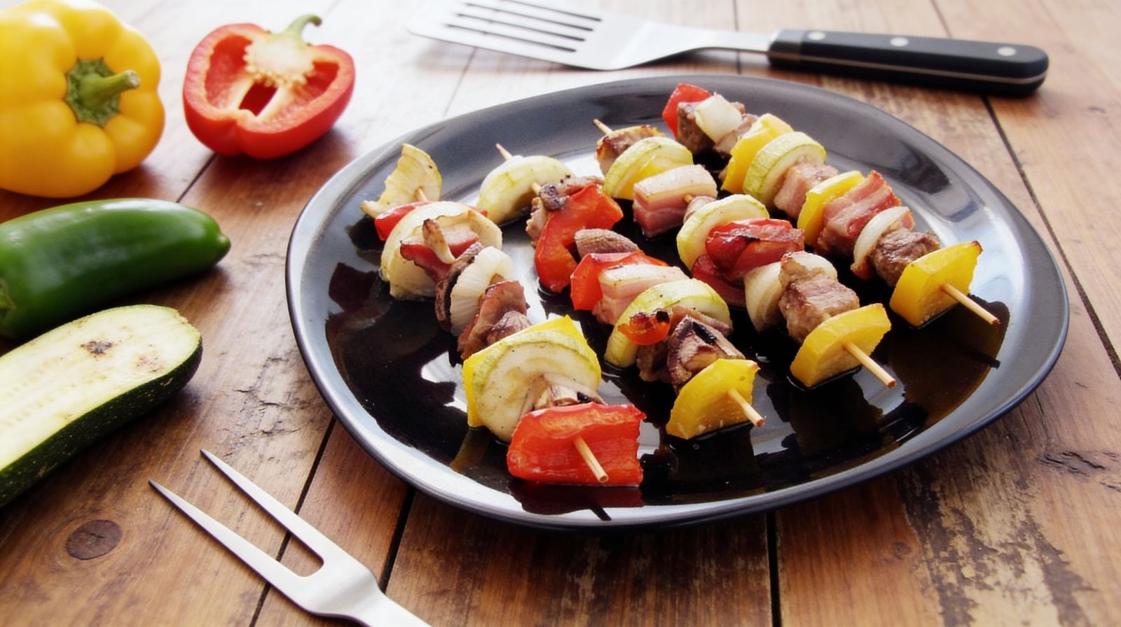 Szaszłyki mięsno-warzywne na grilla i do piekarnika z karkówką, boczkiem, papryką, cukinią i cebulą na czarnym talerzu.
