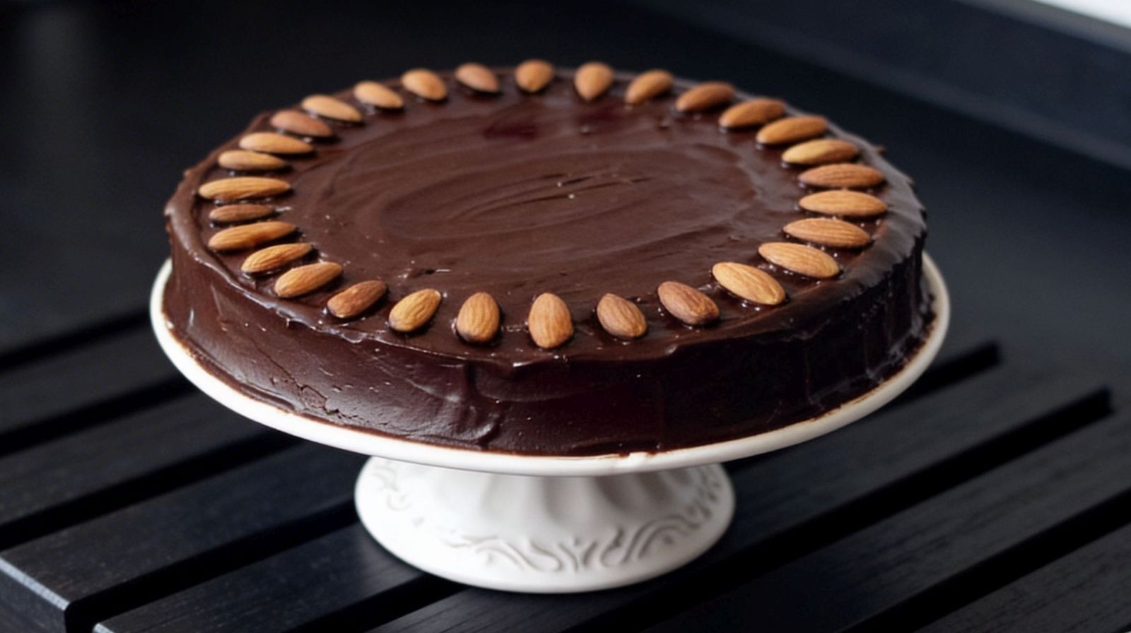 Torcik czekoladowo - migdałowy z polewą z gorzkiej czekolady, udekorowany całymi migdałami, na białej paterze.