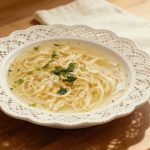 Rosół pieczony klarowny domowy bulion z makaronem nitki i natką pietruszki na białym talerzu.